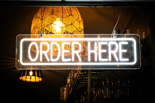 Bright neon 'Order Here' sign illuminated in a cozy café ambiance with warm lighting.